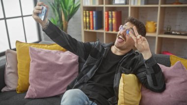 Neşeli bir İspanyol adam, sıcak bir oturma odasında barış işaretiyle selfie çekiyor..