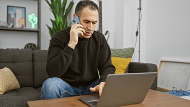 Modern oturma odasında dizüstü bilgisayar kullanan İspanyol bir adam telefonda konuşuyor.