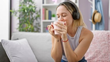 Genç sarışın kadın müzik dinliyor kahve içiyor evde gülümsüyor.