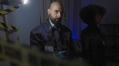 Dedektiflik rozeti takan sakallı bir adam ve kıvırcık saçlı bir kadın, kapalı alanda hafif aydınlatılmış bir suç mahallini dikkatli bir bantla araştırıyor..