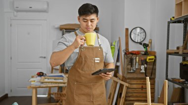 Önlüklü Asyalı adam kahve içer ve aletlerle dolu bir atölyede tablet kullanır..