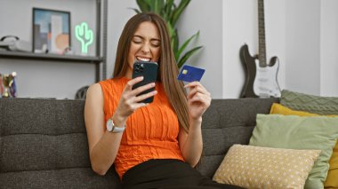 Akıllı telefon kullanan ve elinde kredi kartı olan neşeli bir kadın modern bir oturma odasında online alışveriş neşesini dile getiriyor..