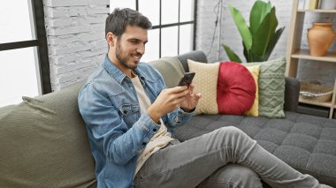 Yakışıklı İspanyol adam, modern ev dekorasyonuyla kanepede akıllı telefon kullanıyor.