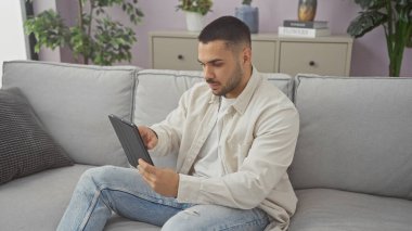 Yakışıklı İspanyol adam, rahat bir oturma odasında gri bir kanepede tablet kullanıyor..