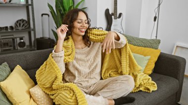 Evinde sarı bir battaniyeyle kanepede dinlenen gülümseyen bir kadın. Sıcak ve pozitif bir hava yayıyor..