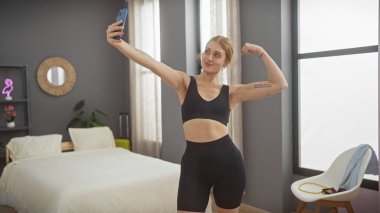 Kafkasyalı genç kadın rahat bir yatak odasında kaslarını esnetirken özgüvenli ve zinde bir şekilde selfie çekiyor..