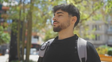 Şehir arka planında bulanık bir şehir parkında dalgın dalgın duran sakallı genç bir adam.