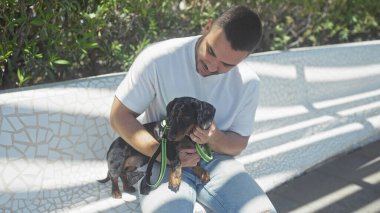 Genç bir İspanyol adam güneşli bir şehir parkında bankta otururken şefkatle bir daksund köpeği tutuyor..