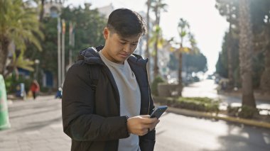 Akıllı telefon kullanan Asyalı genç bir adam palmiye ağaçlarıyla kaplı güneşli bir caddede duruyor..