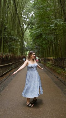 Gözlüklü neşeli İspanyol kadın dönüyor, güzel elbisesi Kyoto 'nun büyüleyici bambu ormanında akıyor.