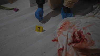 Bir adam kanlı bir suç mahallini kanıt numaraları ve adli aletlerle evin içinde inceliyor..