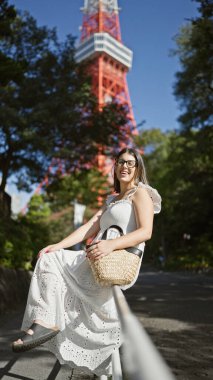 Beautiful hispanic woman in glasses happily enjoying her vacation, smiling by tokyo's famous tower, sky-high adventure and cityscape delight in tokyo clipart