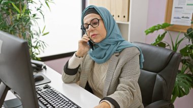 Professional middle-aged woman in hijab speaking on phone in modern office setting clipart