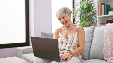 Gülümseyen, gri saçlı, rahat bir oturma odasındaki kanepede dizüstü bilgisayar kullanan olgun bir kadın..