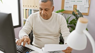 Ofisinde çalışan olgun bir İspanyol adam bilgisayar kullanıyor ve belgeleri inceliyor..