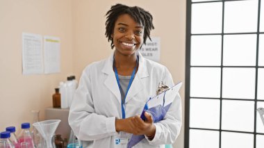 Laboratuvar önlüklü gülümseyen Afrikalı Amerikalı kadın laboratuvar ortamında elinde bir pano tutuyordu..