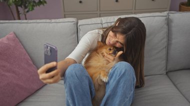 Rahat bir oturma odasında genç bir kadın akıllı telefonuyla selfie çekerken Pomeranian köpeğine sevgiyle sarılıyor..