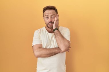 Sarı arka planda duran sakallı orta yaşlı bir adam kollarını kavuşturarak depresyondan sıkılmış ve yorgun göründüğünü düşünüyor.. 