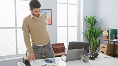 Modern bir ofiste dizüstü bilgisayarı, kulaklığı ve gün ışığıyla düşünceli bir şekilde duran İspanyol bir adam..