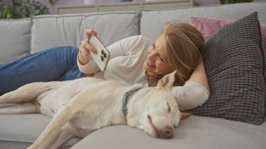 Evdeki kanepede uzanan genç bir kadın akıllı telefonunu kullanırken köpeğiyle vakit geçiriyor..
