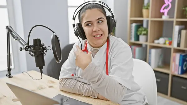 stock image A smiling young woman wearing headphones sits in a modern radio studio, gesturing while recording a podcast.