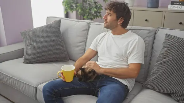 A young hispanic man relaxes at home on a couch, holding a coffee cup and petting a siamese cat.