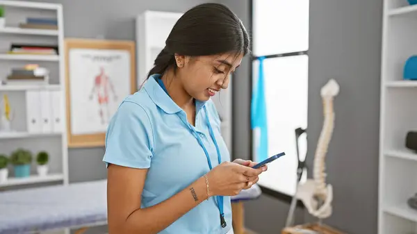 stock image An indian woman in a clinic engages with her smartphone amidst medical equipment, conveying modern healthcare multitasking.