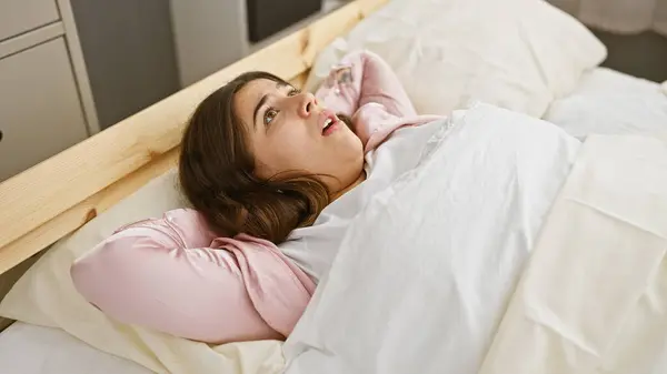 stock image Young hispanic woman, looking worried while lying in bed inside a bedroom with neutral-colored bedding and minimal decor.