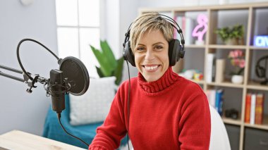 Kısa sarı saçlı, gülümseyen İspanyol kadın radyo stüdyosunda kulaklık takıyor.