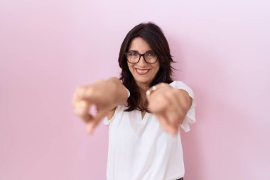 Orta yaşlı, beyaz tişörtlü ve gözlüklü İspanyol kadın parmaklarıyla seni ve kamerayı işaret ediyor. 