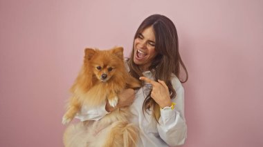Neşeli genç bir kadın pembe izole edilmiş arka planda Pomeranian köpeğini işaret ediyor.