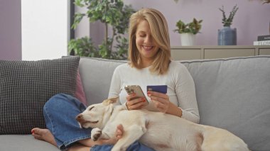 Elinde kredi kartıyla akıllı telefon kullanan, rahat bir oturma odasında bir köpekle koltukta uzanan gülümseyen bir kadın..
