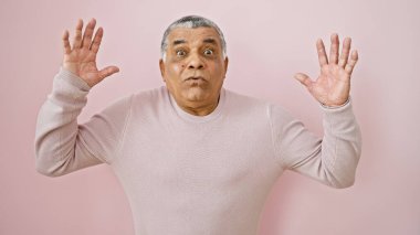 Şaşırmış orta yaşlı, gri saçlı, pembe arka planda el kaldıran bir adam.