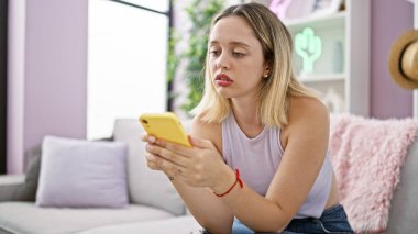 Evdeki kanepede oturan akıllı telefon kullanan genç sarışın kadın.