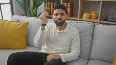 Düşünceli, sakallı genç bir İspanyol adam modern bir oturma odasında bir koltukta oturur ve tefekkür eder.