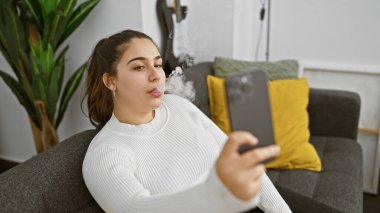 İçerde buhar yapan genç İspanyol kadın, rahat bir oturma odasında telefonunu kullanıyor..