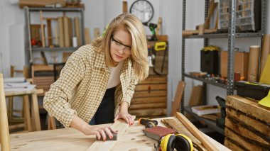 Sarışın bir kadın atölyede ahşap zımparalıyor, işçilik ve yaratıcı stüdyo çalışmaları çağrıştırıyor..