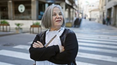 Gri saçlı, düşünceli yaşlı bir kadın şehir sokaklarında çapraz kollar arasında duruyor, şehir zarafetini somutlaştırıyor..