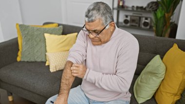 Olgun, kır saçlı bir adam, kanepede otururken kolunu kaşıyor.