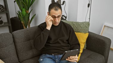 Kapalı bir koltukta otururken elinde tablet tutan, gözlüklü, düşünceli İspanyol bir adam..