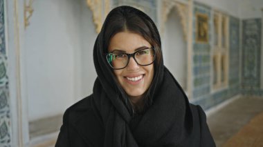 Geleneksel bir Türk iç mekanında siyah başörtüsü takan gözlüklü, gülümseyen bir kadın..