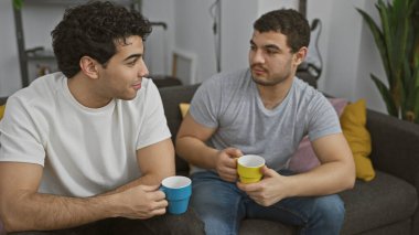 İki adam kapalı bir salonda gri bir kanepede oturuyor, ellerinde renkli kupalar, sıcak bir oturma odasında dostça bir sohbete girişiyorlar..