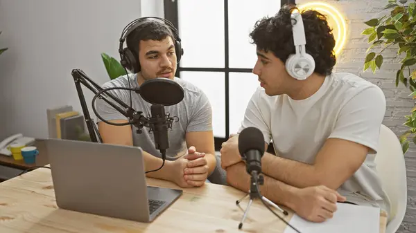 stock image Two men engaged in podcasting indoors, equipped with microphone, headphones, laptop, and a studio setting.