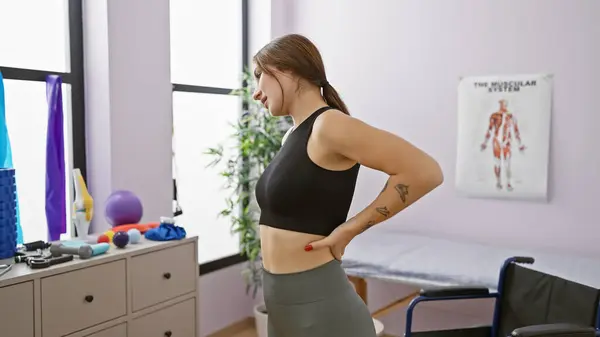 stock image A young caucasian woman with a tattoo stands thoughtfully in a rehabilitation clinic, surrounded by exercise equipment.