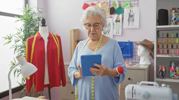 stock image Senior woman tailor with glasses and measuring tape using tablet in colorful atelier.