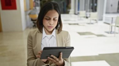 Profesyonel kıyafetli, odaklanmış bir İspanyol kadın modern dekorlu bir ofis koridorunda tablet kullanıyor..