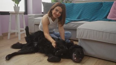 Şık bir oturma odasında genç bir kadın, mutlu labrador köpeğini yerde, rahat gri bir kanepe ve mavi bir battaniyenin yanında okşar..
