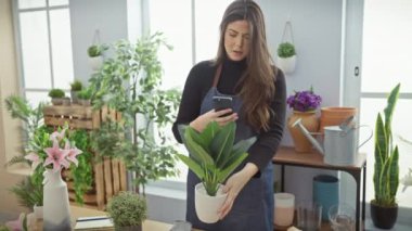 Genç bir kadın akıllı telefonunu kapalı bir çiçekçide bitkilerin fotoğraflarını çekmek için kullanıyor, gelişigüzel ve yeşil bir ortam sergiliyor..