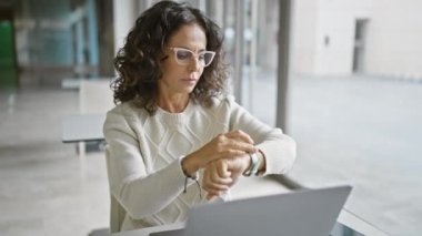 Profesyonel bir kadın modern bir ofis ortamında dizüstü bilgisayarda çalışırken saatini kontrol eder..