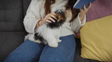 Rahat bir kazak giyen bir kadın renkli yastıklı bir kanepede bisiklet teriyerinin yanında kitap okur..